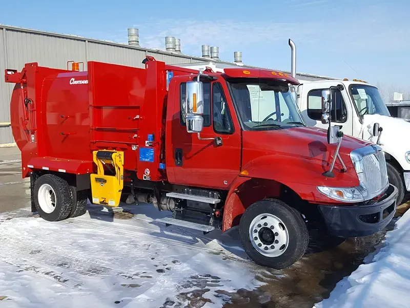 Red garbage truck parked on a snowy street beside white utility vehicles