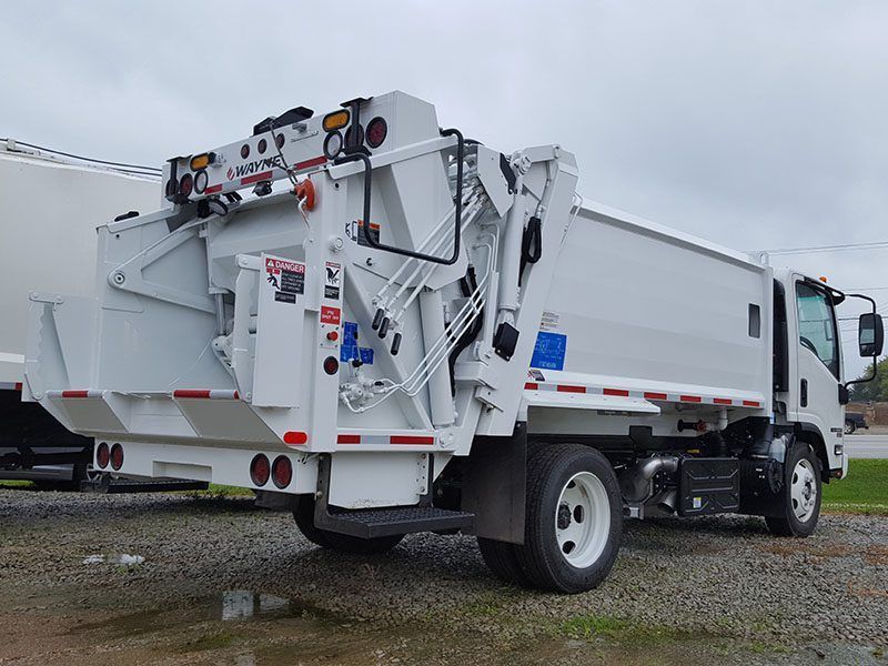 White garbage truck with rear loading lift parked on a gravel lot under cloudy skies