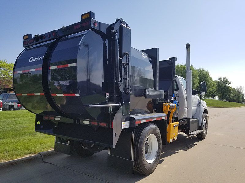 White garbage truck with a side-loading arm parked on a road beside a grassy area