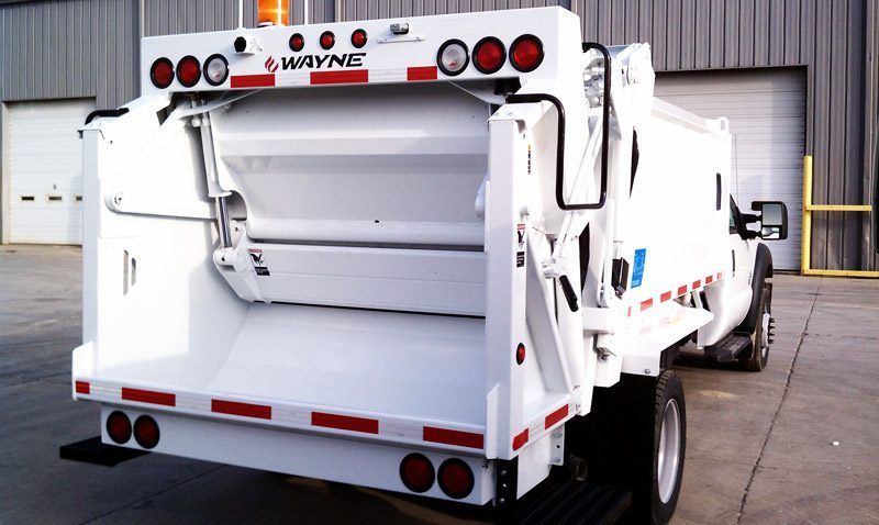 Rear view of a white garbage truck parked outside a warehouse, with an open hopper and red reflectors.