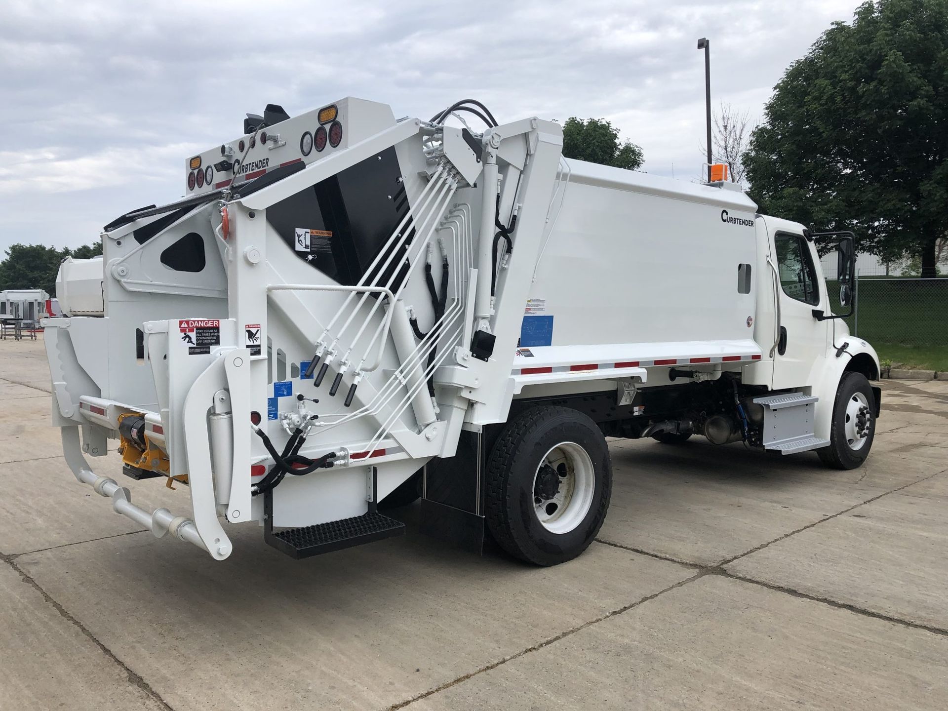 White garbage truck with rear-loading lift parked on a concrete lot under cloudy skies