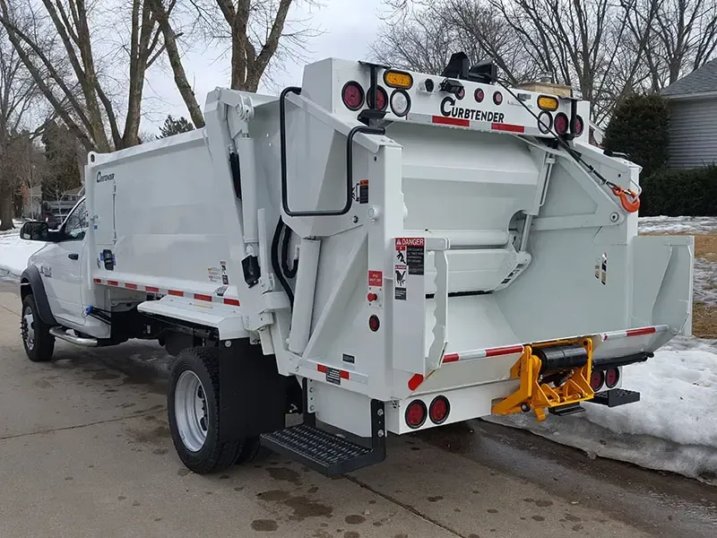 White municipal garbage truck parked on a snowy residential street, rear view.