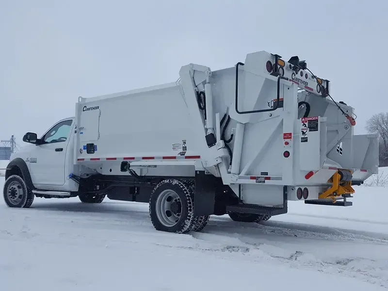 White garbage truck on a snowy road, side view, with rear hopper and snow-covered surroundings