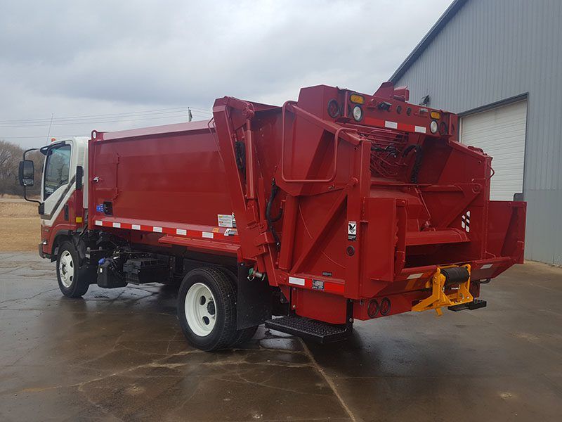 Red garbage truck parked outside on wet pavement near a white garage