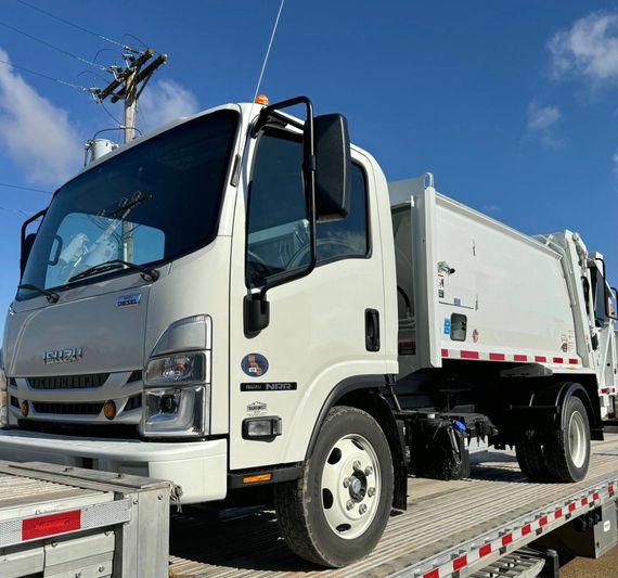 White garbage truck on a flatbed trailer under a clear blue sky.