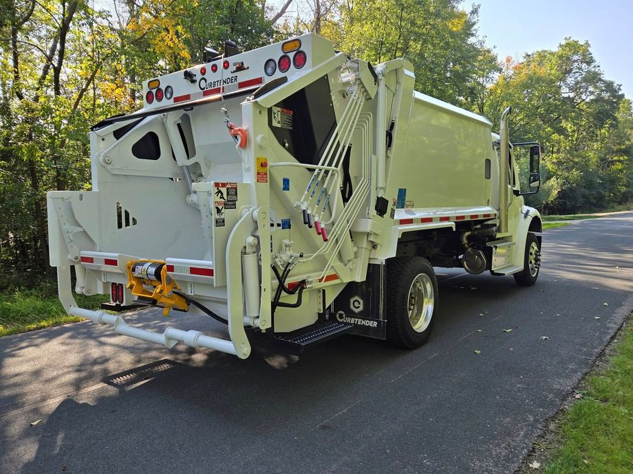 White garbage truck on a tree-lined road, viewed from the rear-left side.