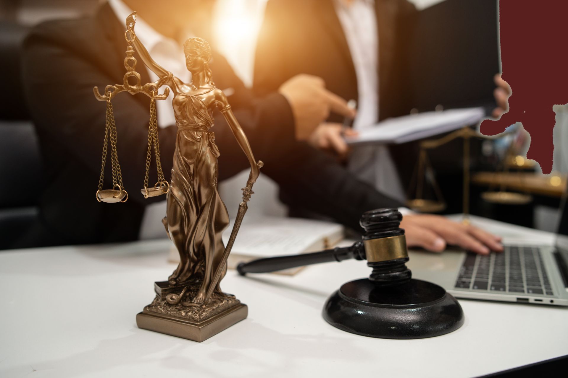 Statue of Lady Justice with scales and gavel on desk; two people in suits, one using a laptop.
