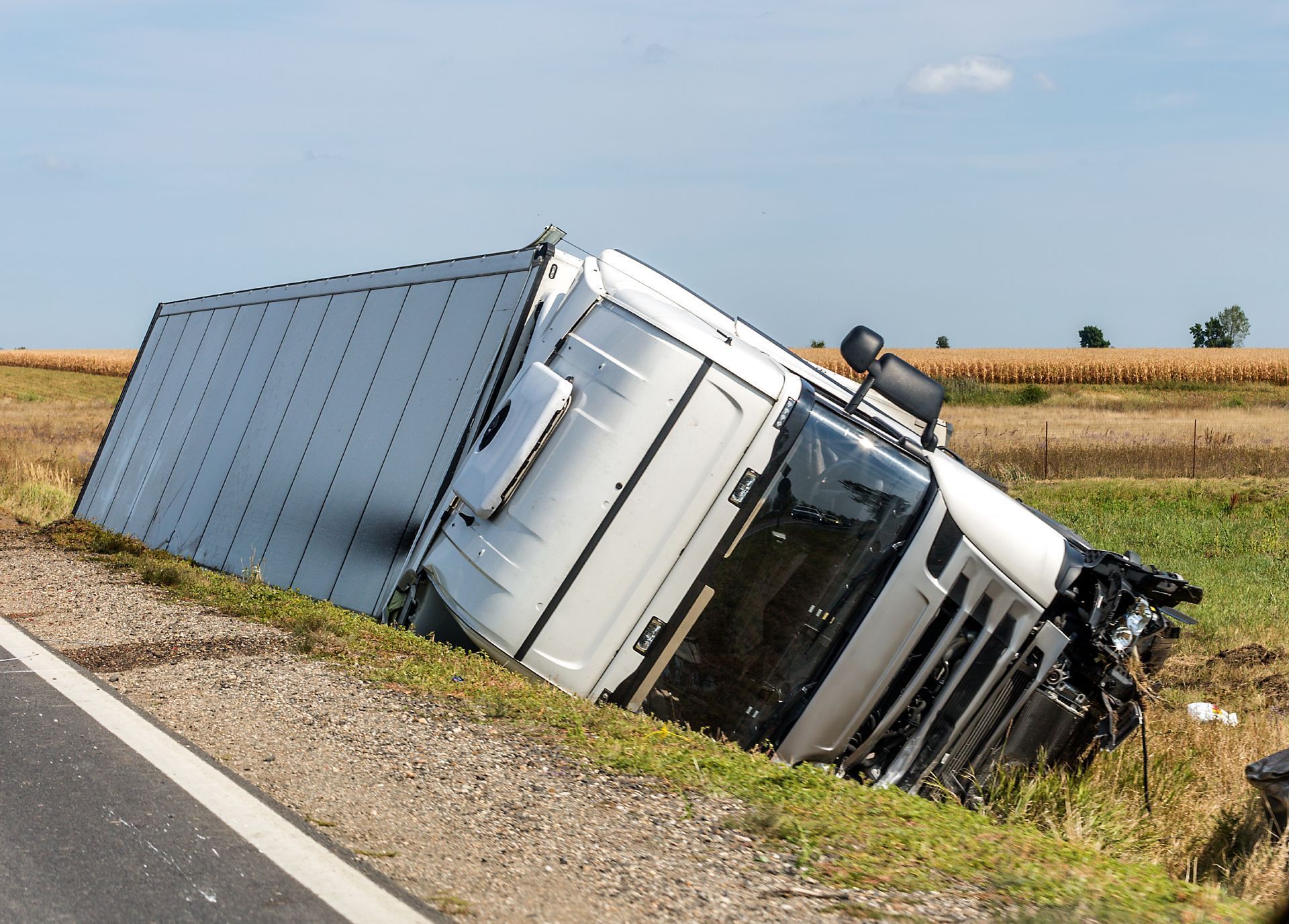 Overturned white semi-truck in a ditch next to a road, field in background.
