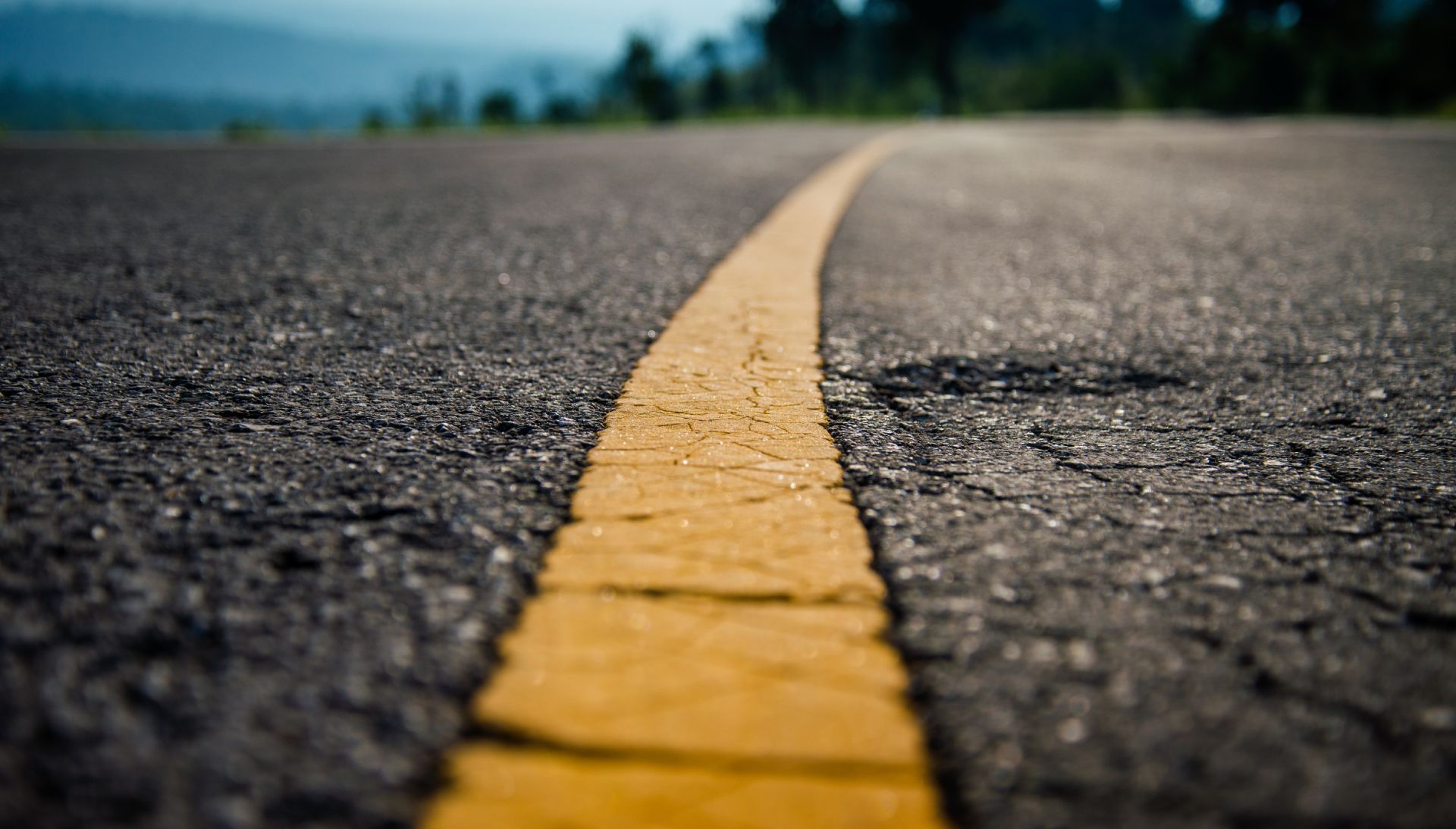 Close-up of asphalt road with yellow dividing line, blurred background of trees and sky.