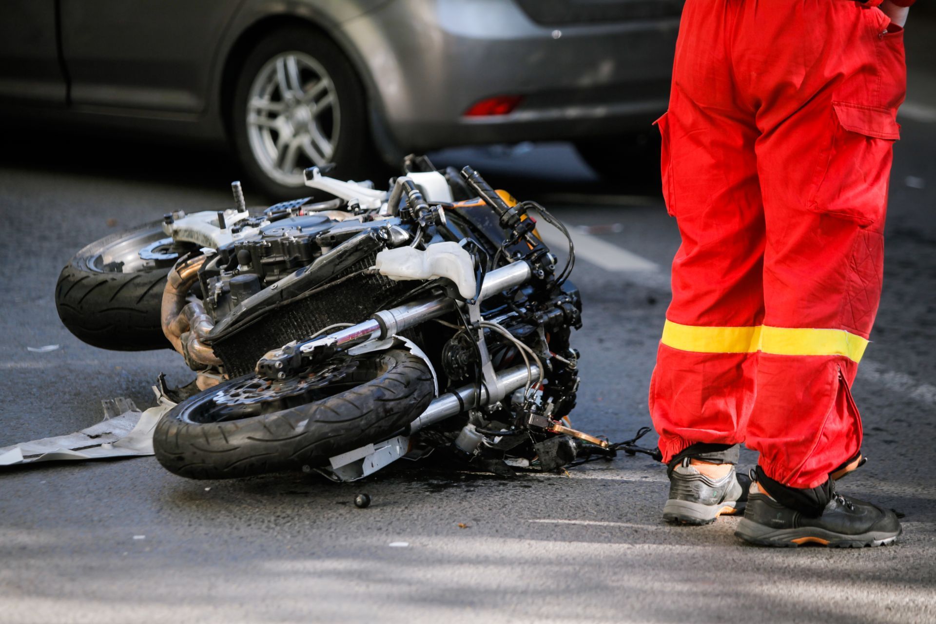 Motorcycle accident: crashed motorcycle on road, emergency responder in red pants standing near a car.