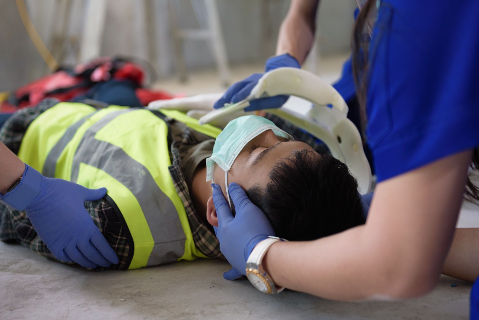 Person on the ground, wearing a neck brace, mask, and reflective vest, receiving medical attention. Two people in blue gloves assist.