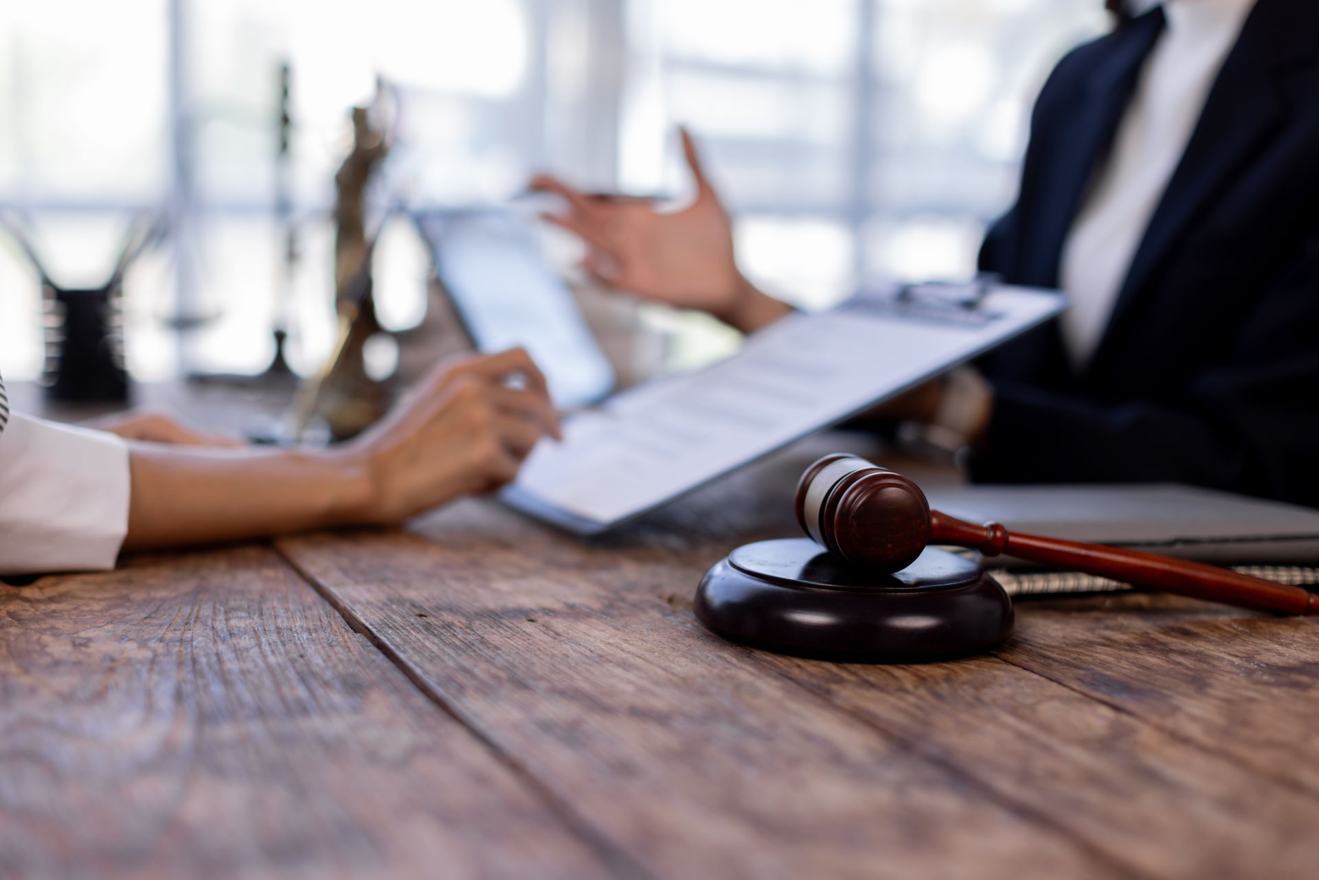 A gavel on a wooden table, with two people discussing documents and a tablet in the background.