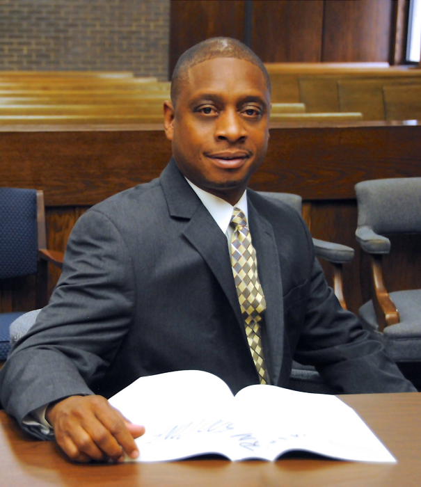 Man in a suit sits at a desk in a courtroom, looking at a document.