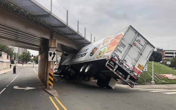 Truck wedged under a low bridge; the trailer is heavily damaged and leaning.