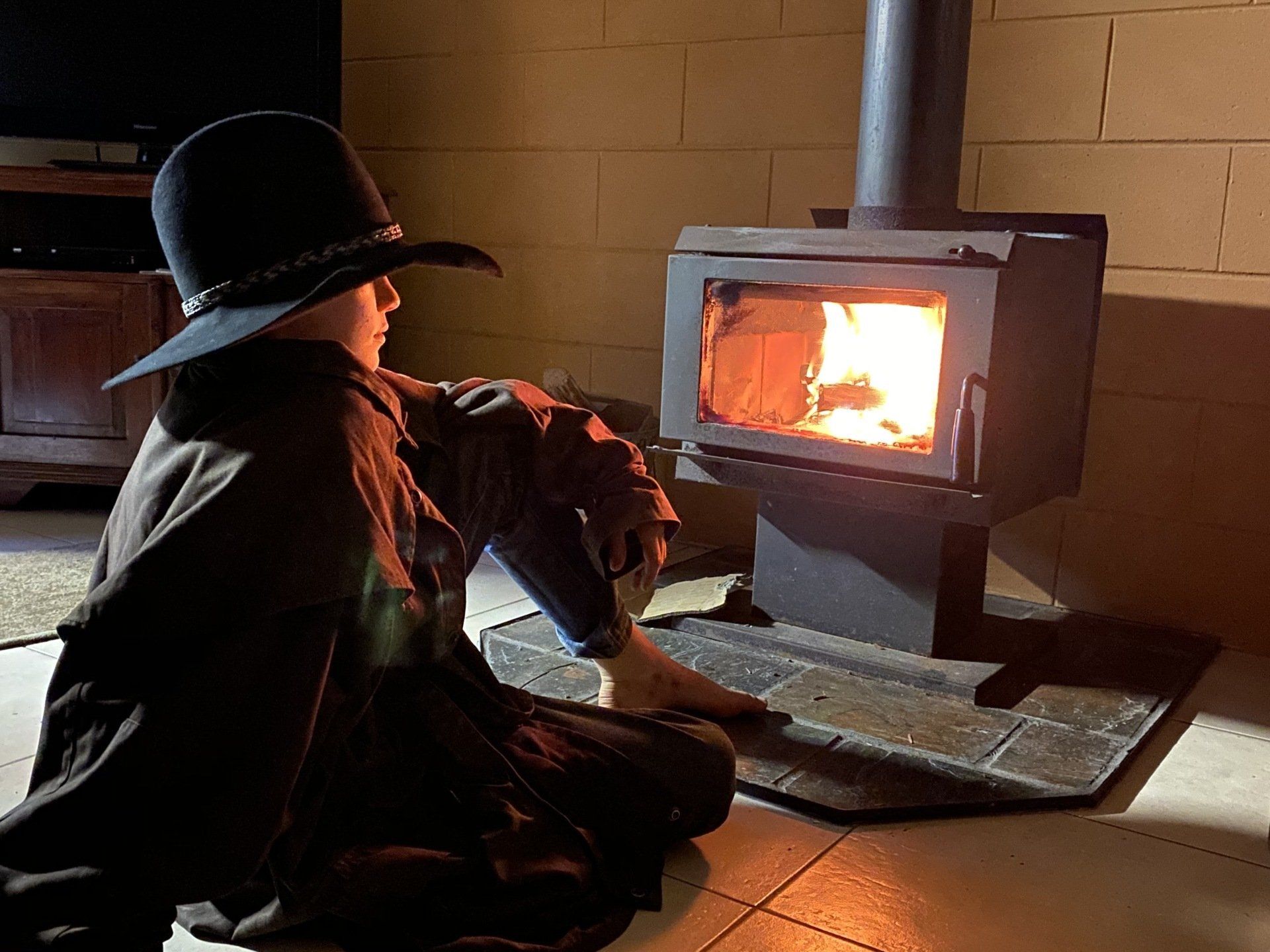 Person in hat and drizabone coat sits near a lit wood stove.
