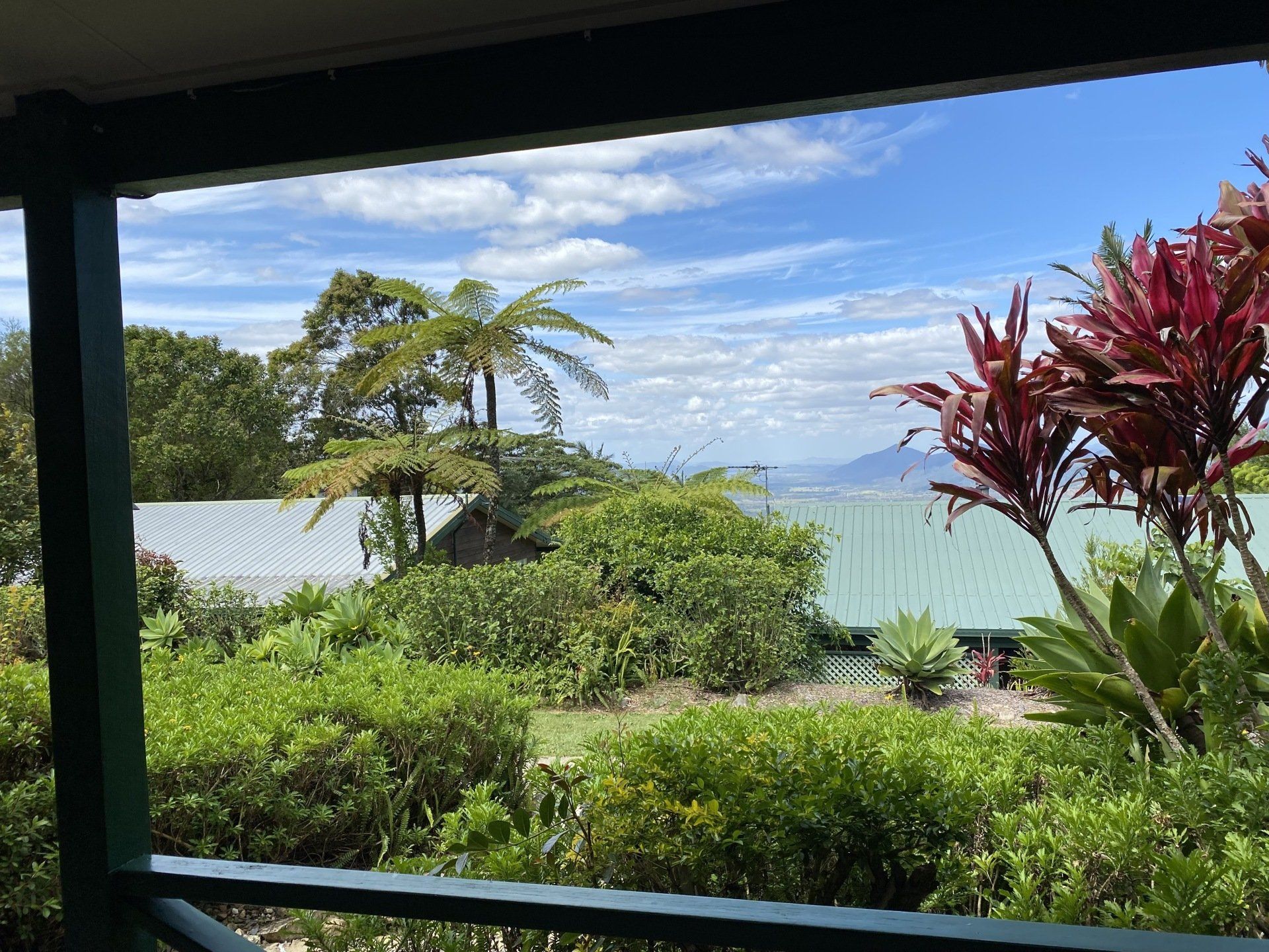 View from a porch with green bushes, trees, and buildings against a blue sky with clouds.