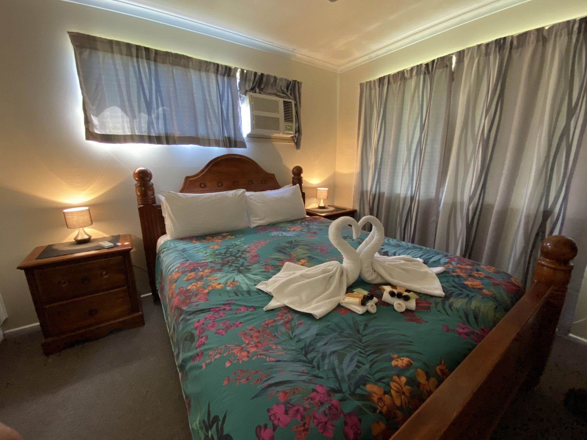 Bedroom with double bed, floral bedspread, swan towel art, nightstands, and window with curtains.