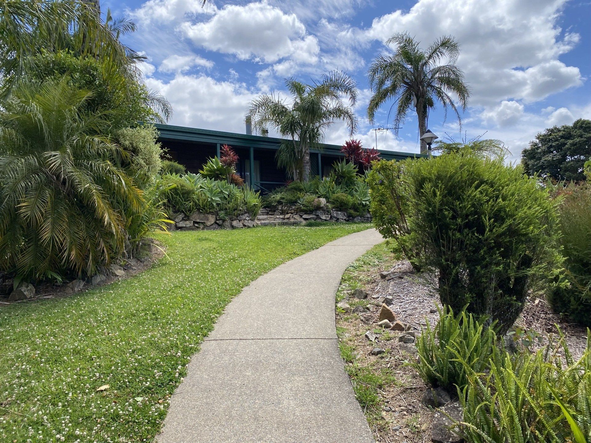 Stone path leads toward a building, surrounded by greenery and trees, under a partly cloudy sky.