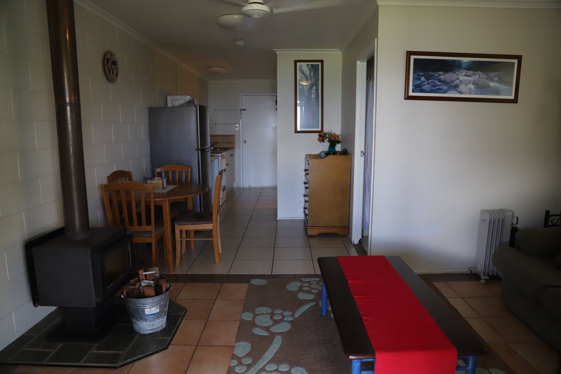 Living room with wood stove, dining table, and sofa.  Kitchen visible in background.  Wooden furniture, neutral walls.