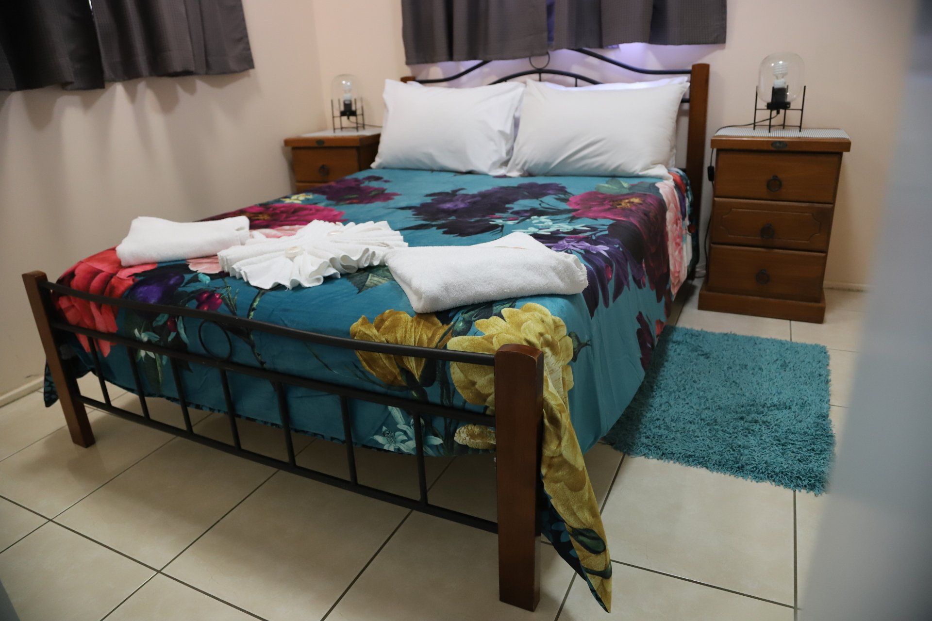 Bedroom in a one bedroom cabin with a made bed featuring a teal floral bedspread, two nightstands, and a blue rug.