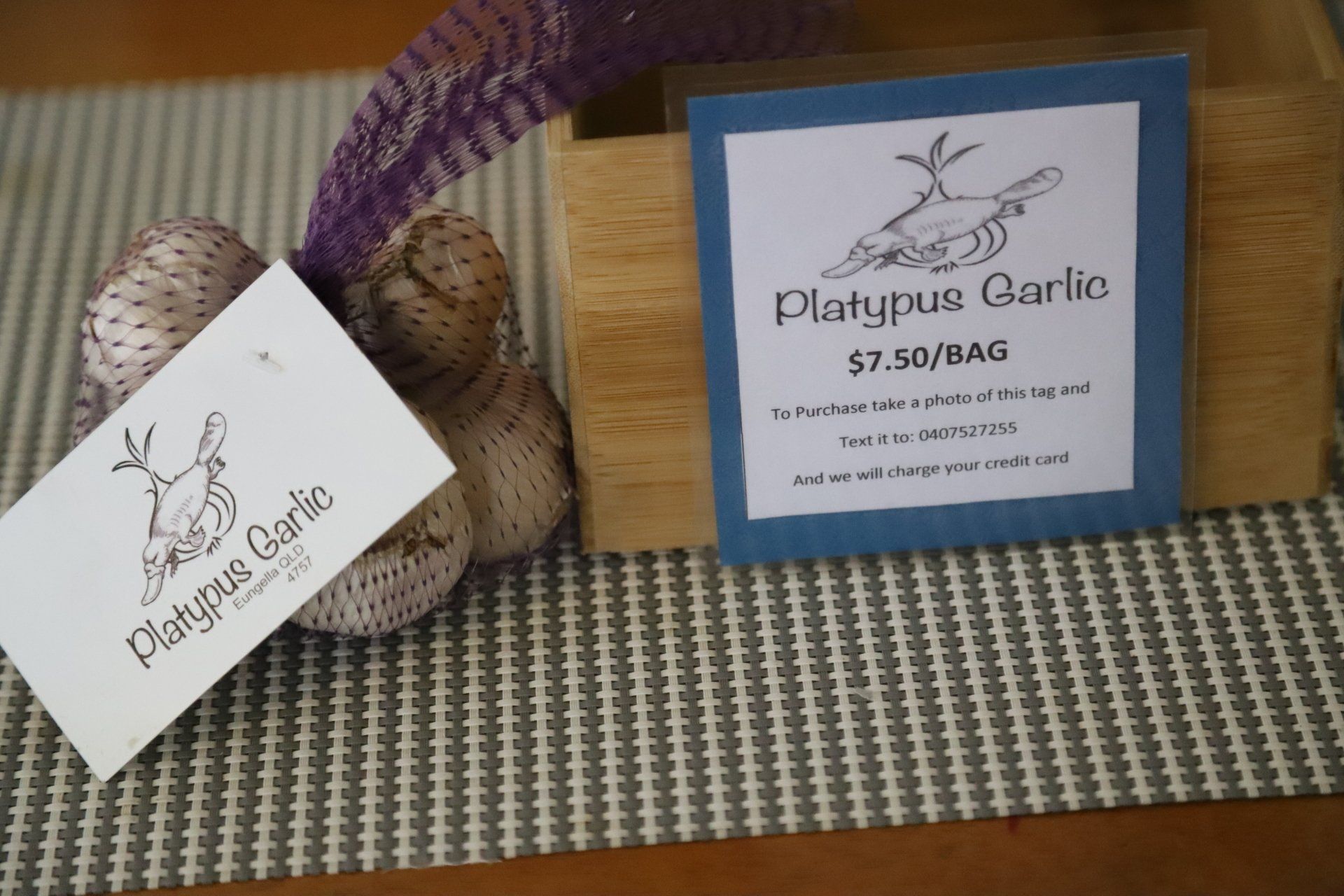 Garlic bulbs in a net bag with a label, and more garlic with a label in a wooden box on a striped placemat.