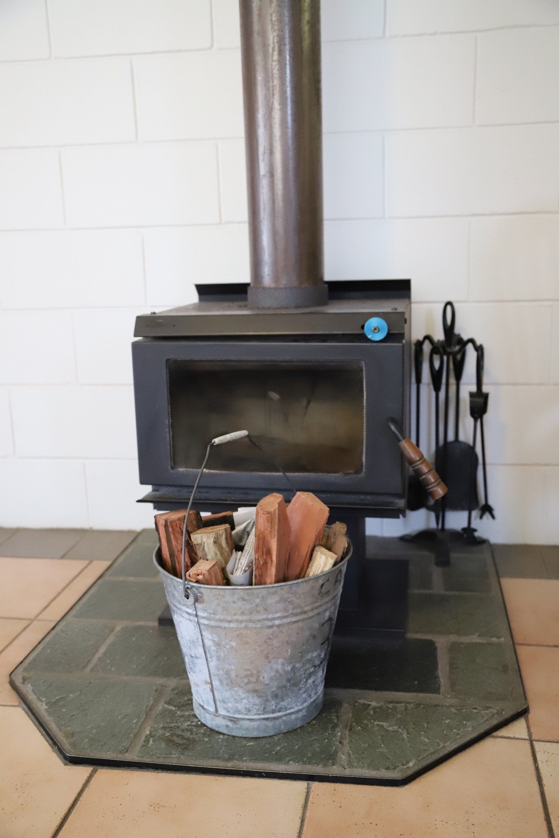 Wood-burning stove with metal bucket of firewood, tools, and a brick wall background.