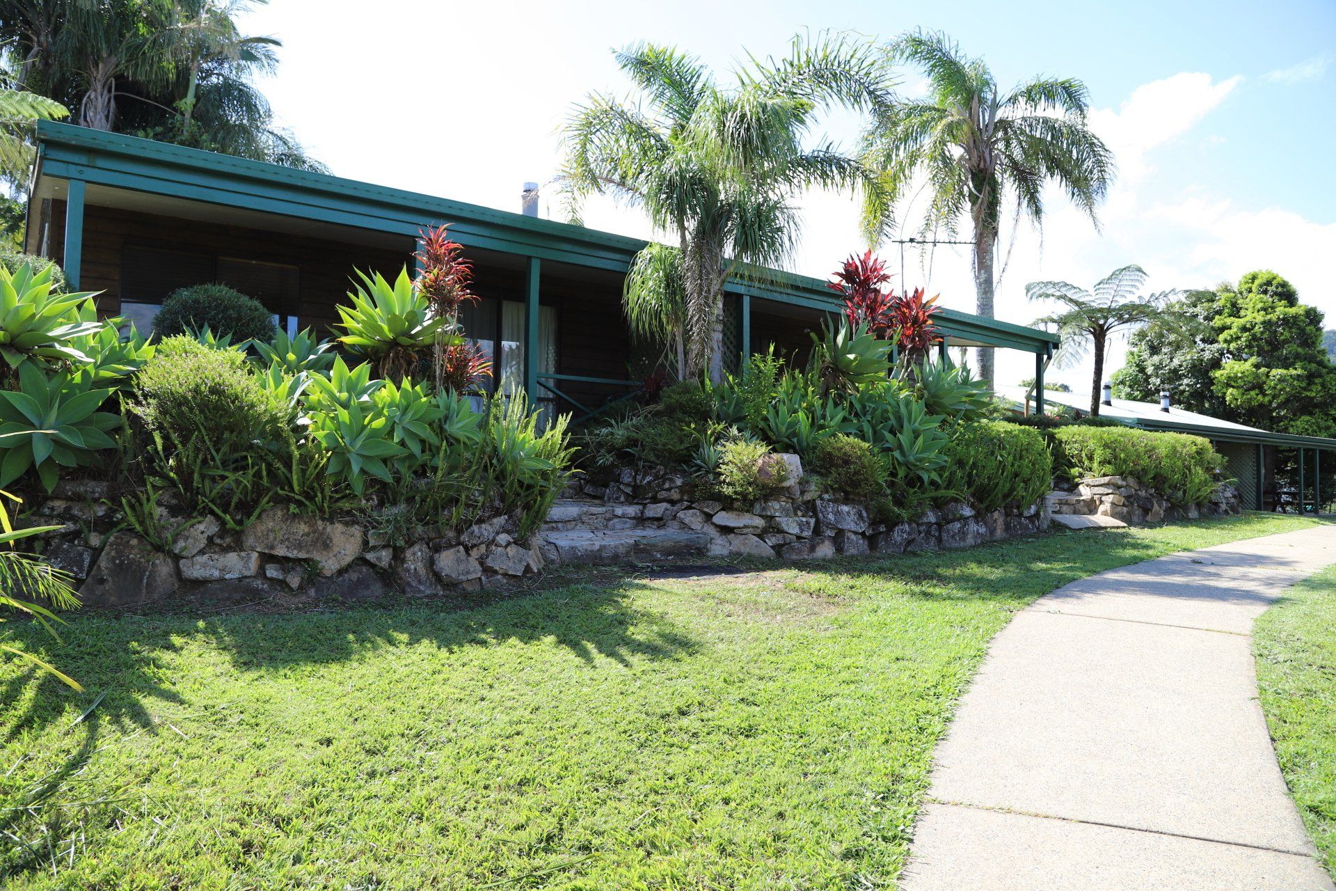 A bungalow-style building with a green roof and lush tropical landscaping. A paved path leads to the structure.