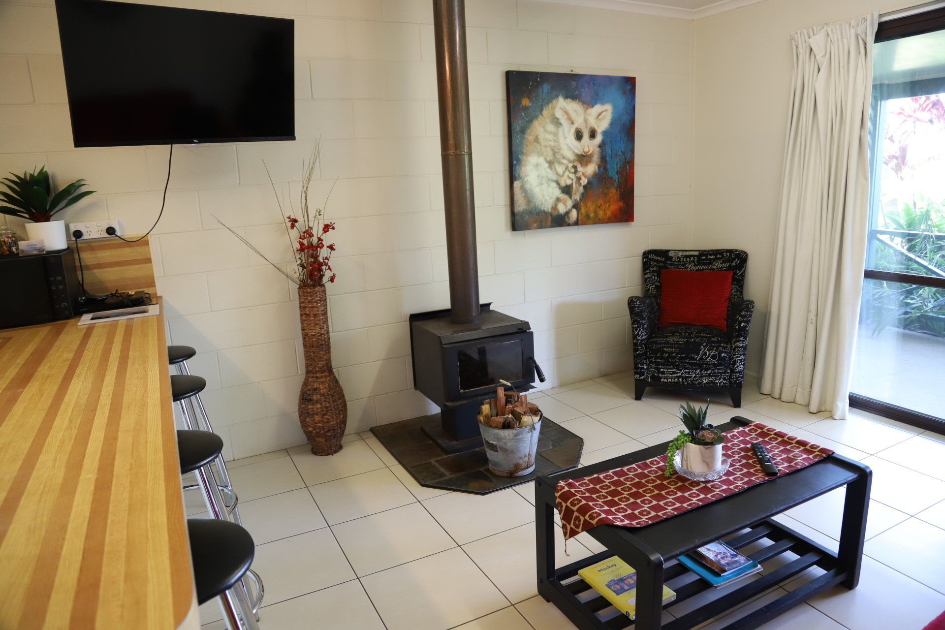 Living room with wood-burning stove, TV, bar stools, armchair, and low table with books and plants, near a sliding door.