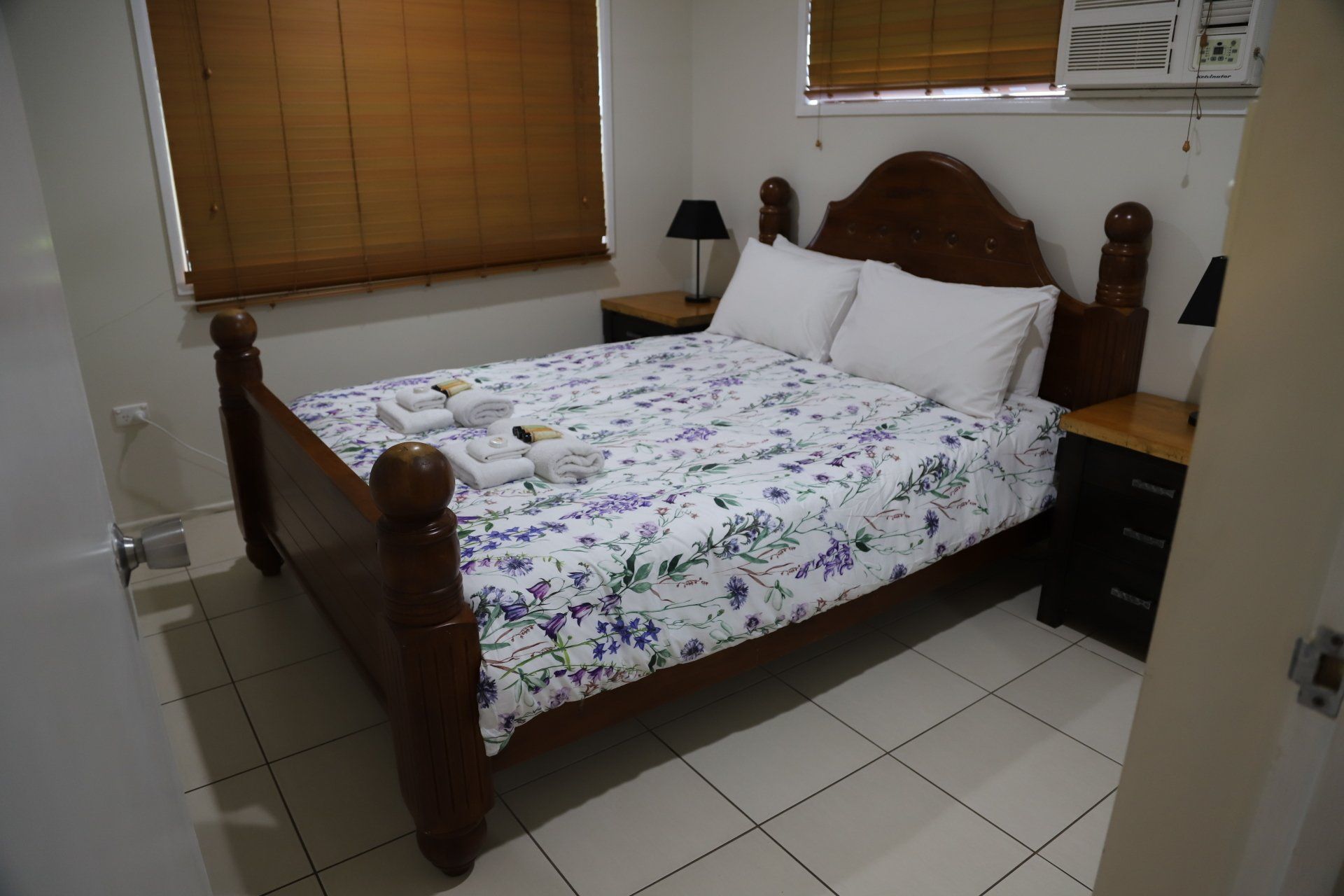 Bedroom with a wooden bed frame, floral bedding, two nightstands, and a window with blinds.