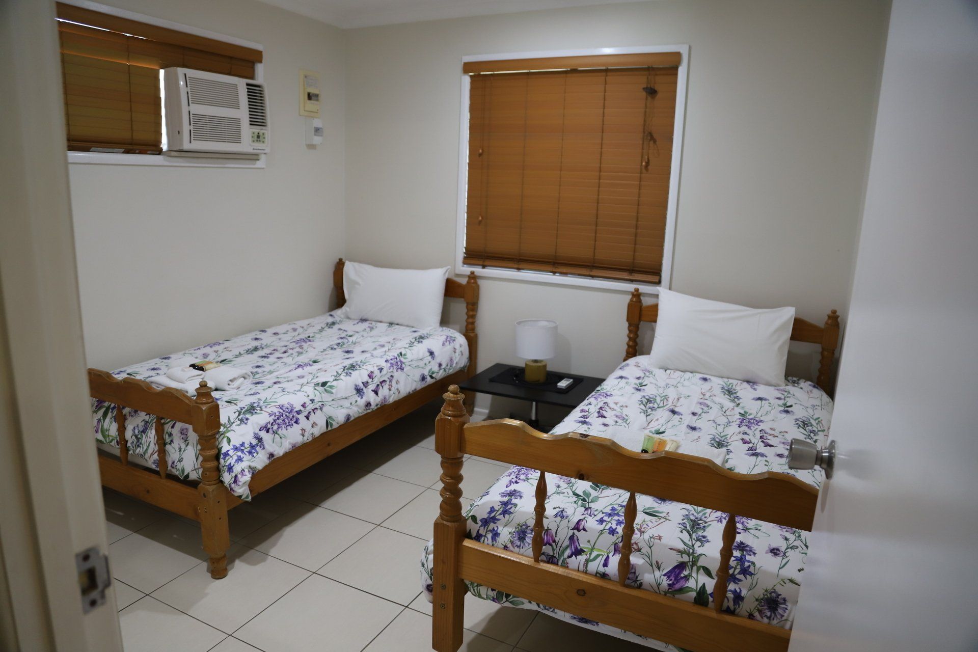 Two twin beds in a room with a window and a small table between them; white walls, patterned bedding.