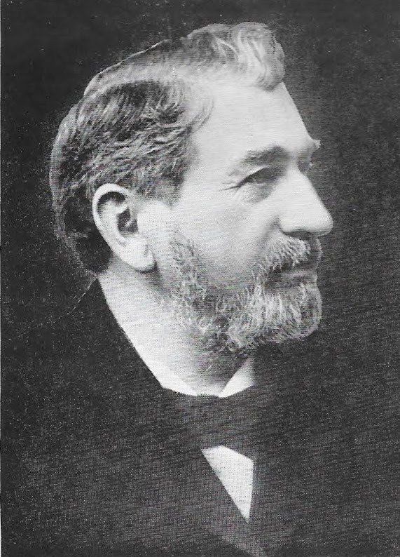 Portrait of a man in a dark suit with a beard, facing right.