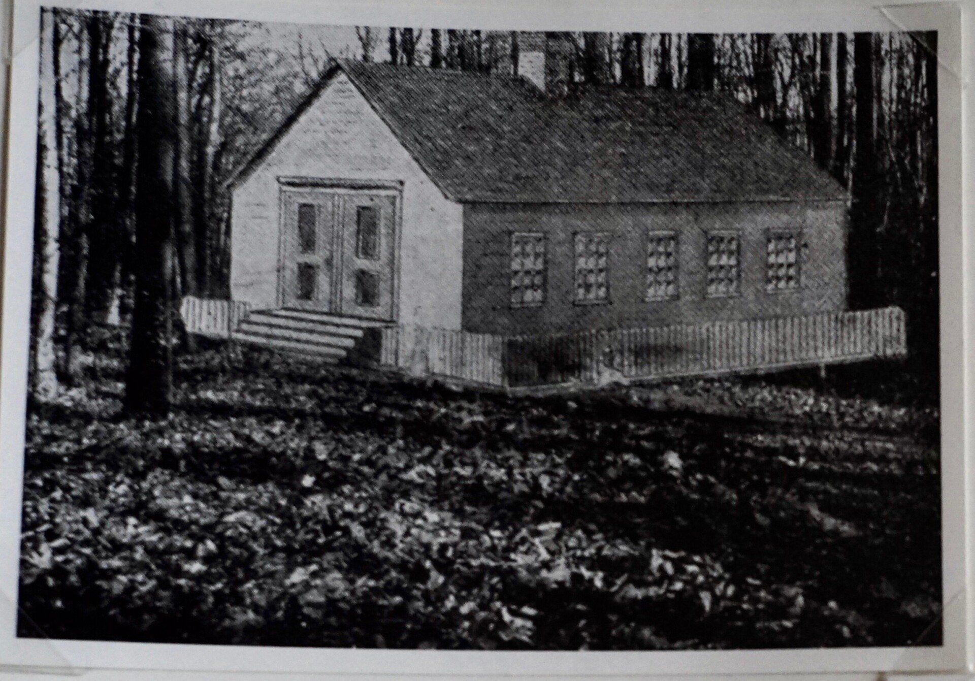 Small, wooden schoolhouse in a forest setting, with a picket fence and steps.
