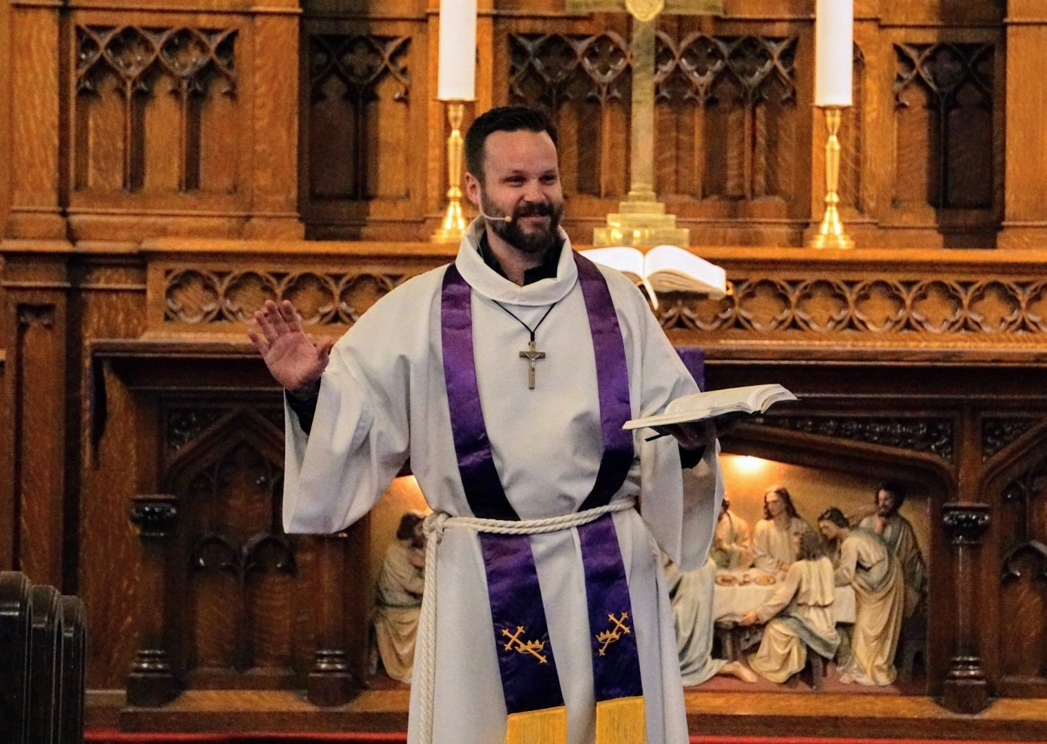 Cleric in white robe and purple stole gestures, holding a book, at church altar with wood paneling.