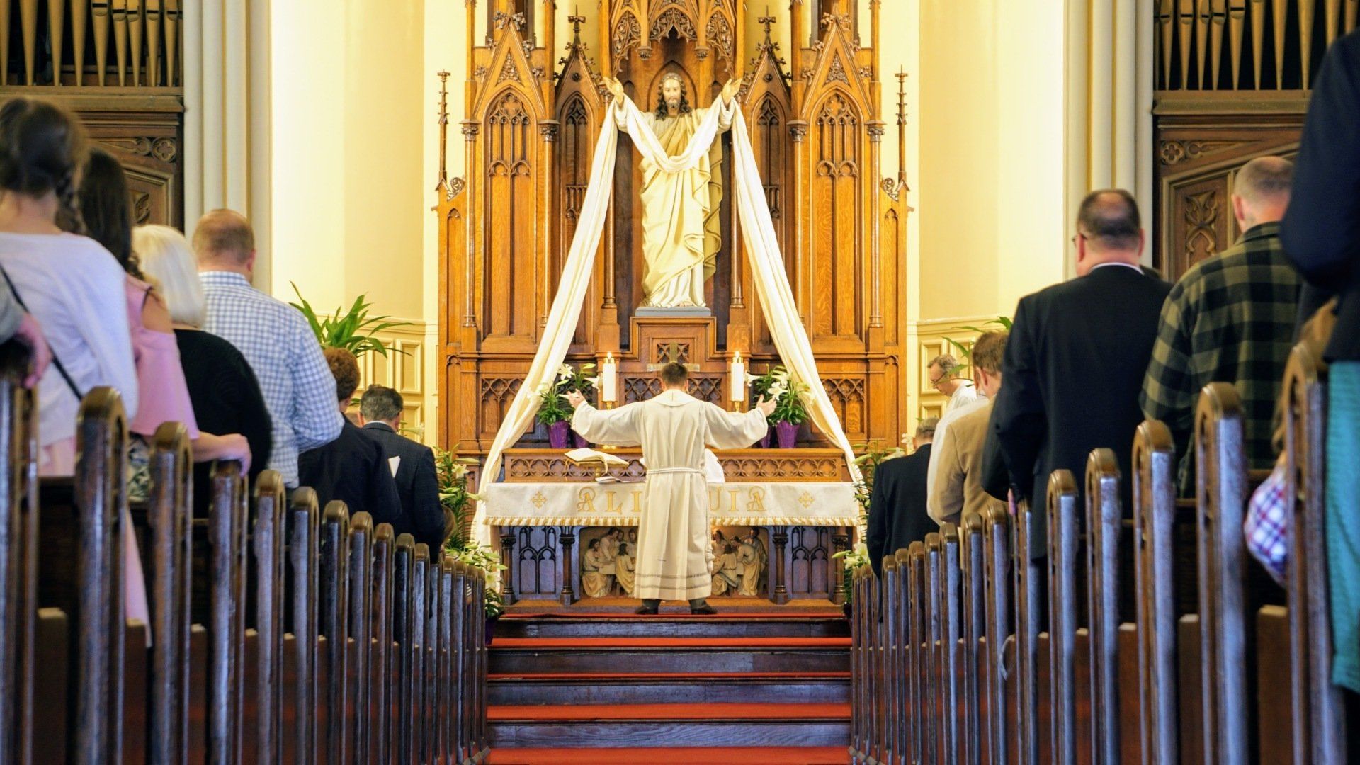 People in a church, facing a priest at an altar, wooden pews, a statue of Jesus in the background.