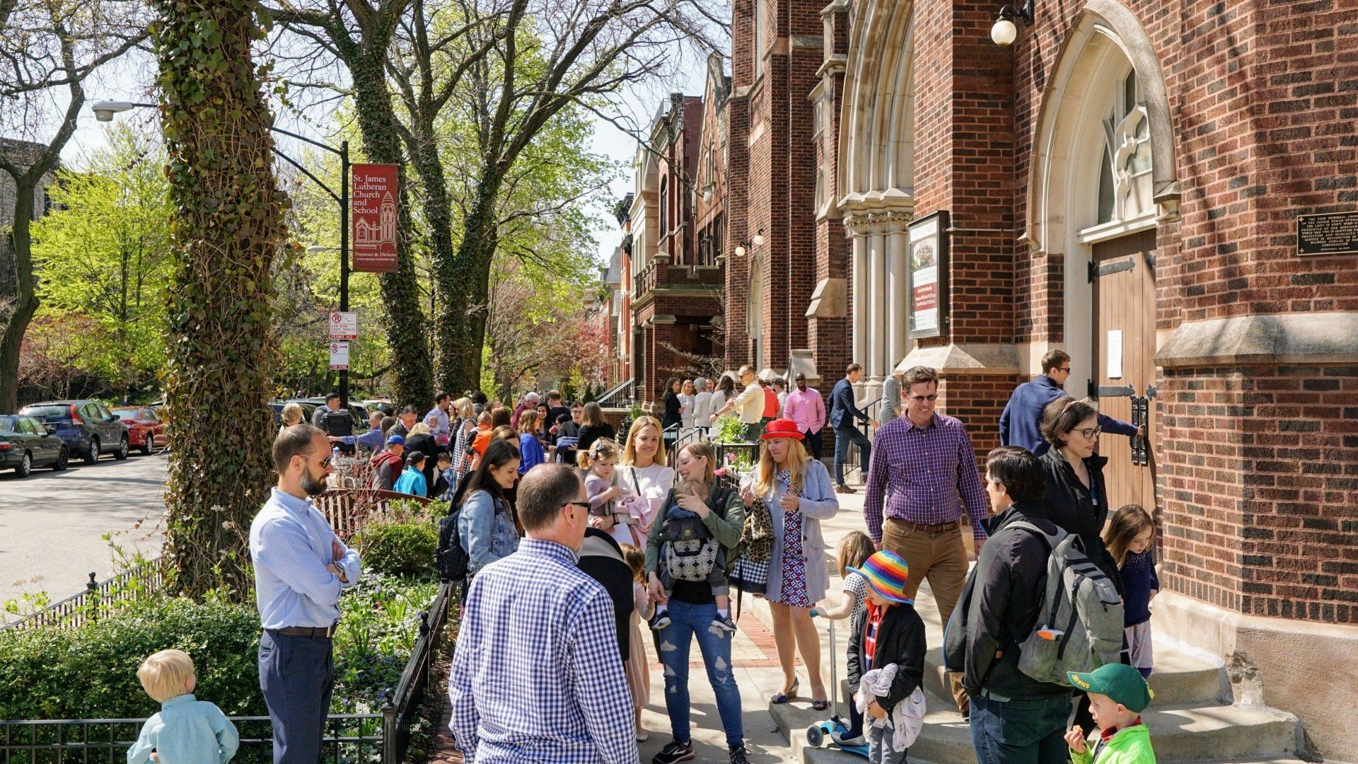 People lined up outside a brick building on a sunny day. Trees and parked cars are visible.
