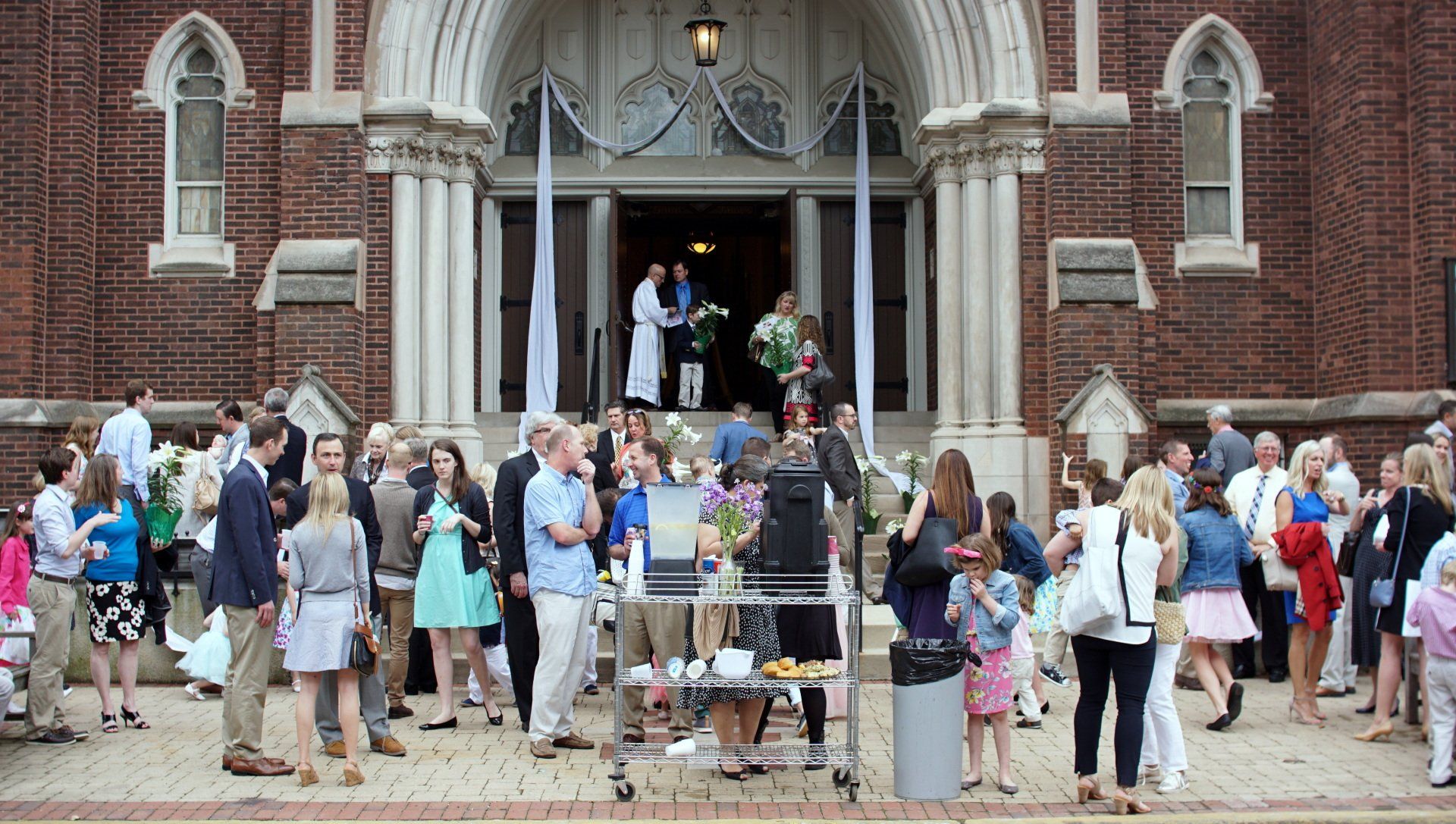 People gathered outside a brick church after a wedding. Some are on the steps, others on the sidewalk.