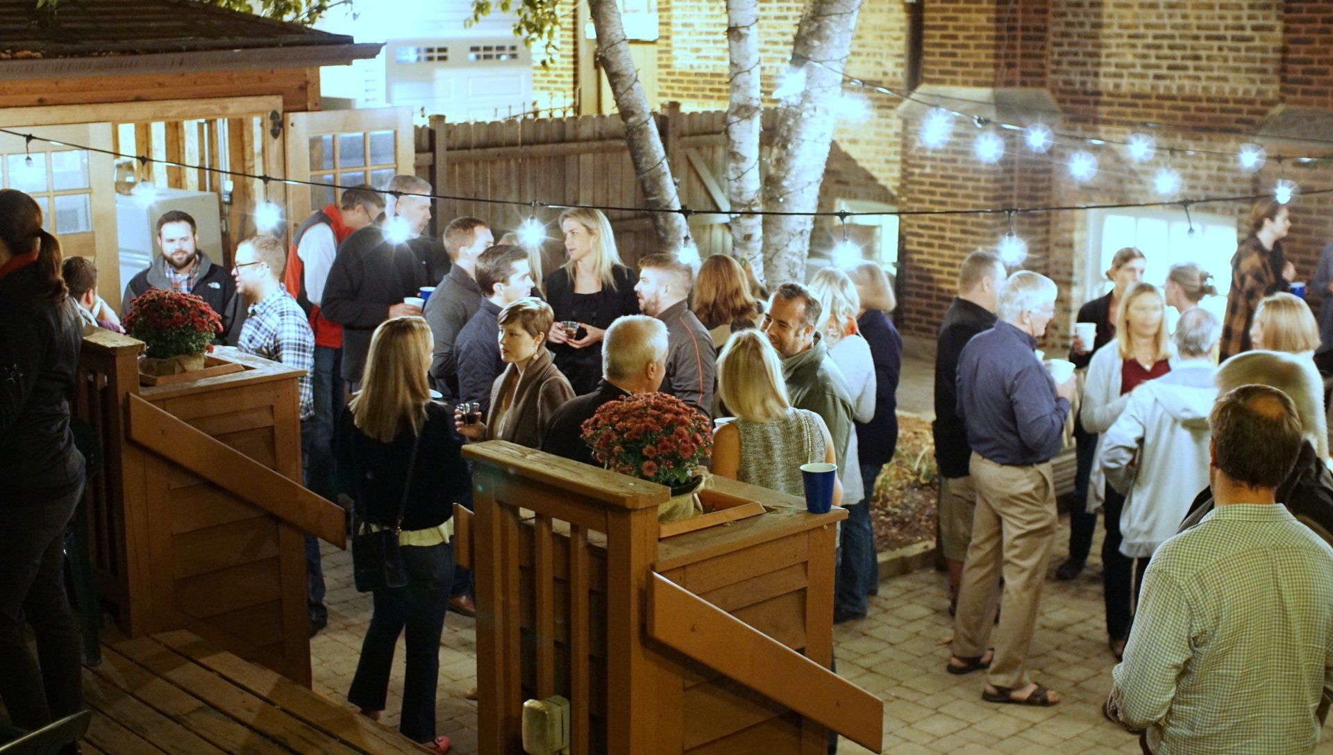 Outdoor gathering with people, string lights, wooden deck, trees, brick building.
