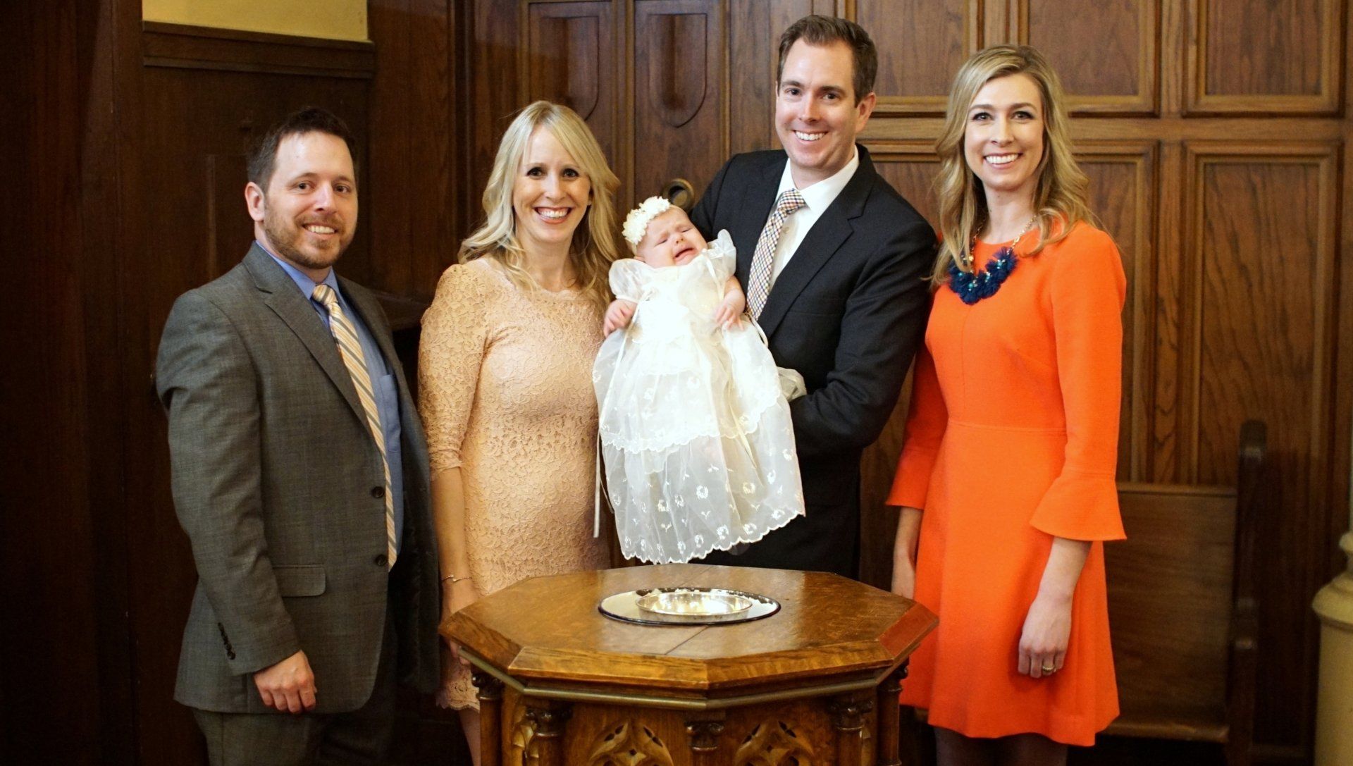 Five people in a church, gathered around a baptismal font with a baby in a baptism gown.
