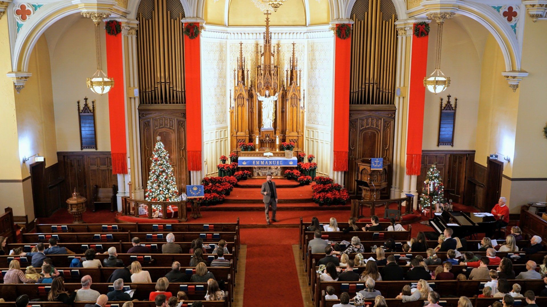 Interior of a church during a service with people seated, red accents, and a speaker at the altar.