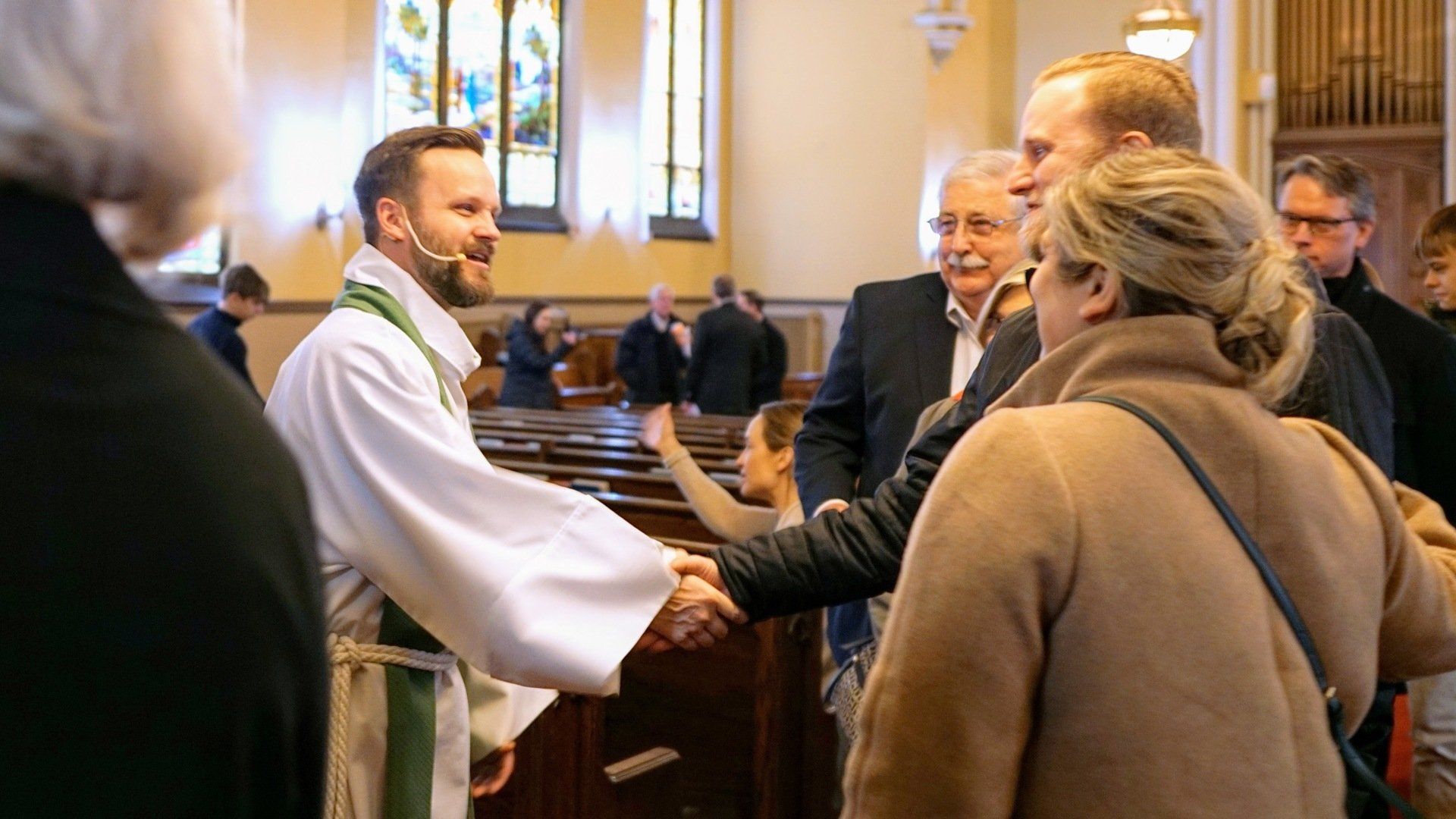 A pastor greets churchgoers. The people are inside a church building. The pastor wears a white robe and smiles.