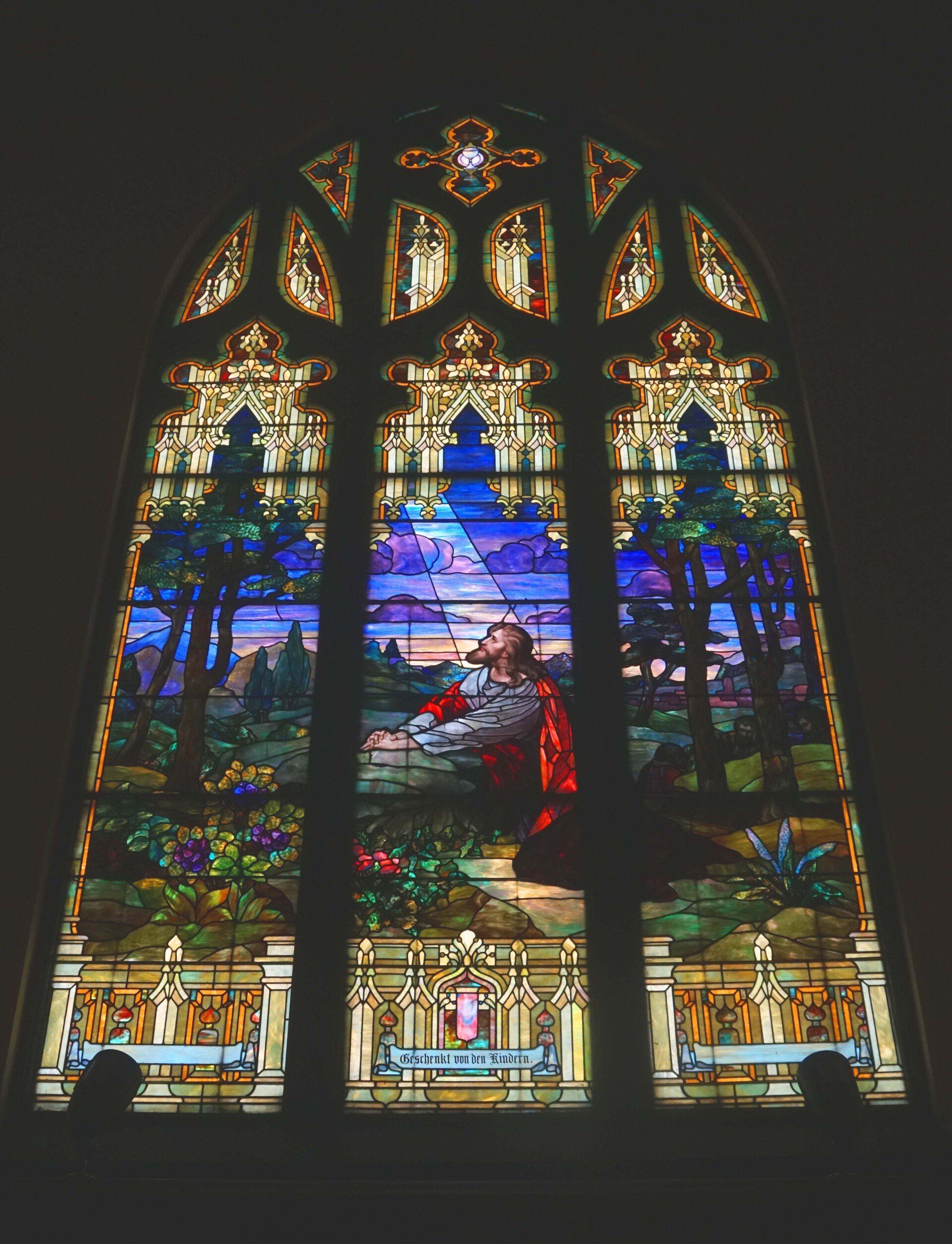 Stained glass window depicting Jesus praying in a garden; vibrant colors and intricate details.