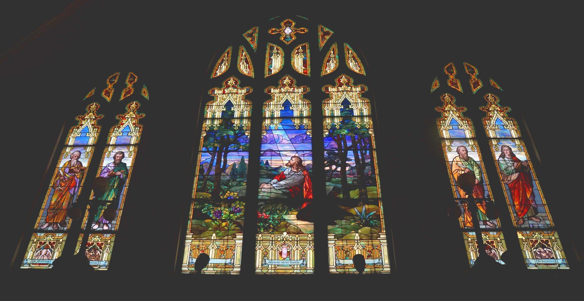 Three ornate stained glass windows with religious figures and landscape in a dark room.