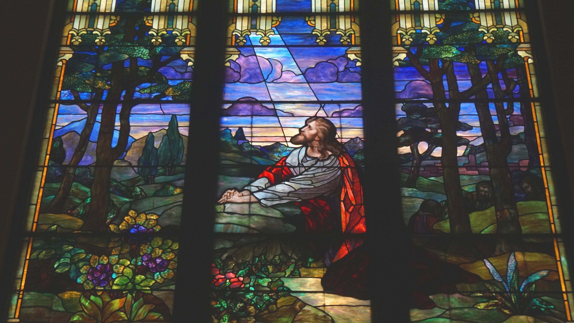 Stained glass window depicts Jesus kneeling in prayer, bathed in light, surrounded by trees and flowers.