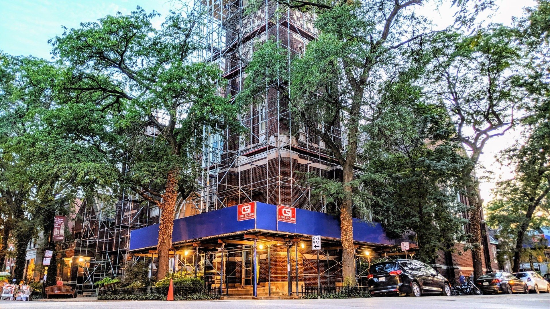 Building facade under construction, surrounded by trees and cars. Blue scaffolding, dusk lighting.