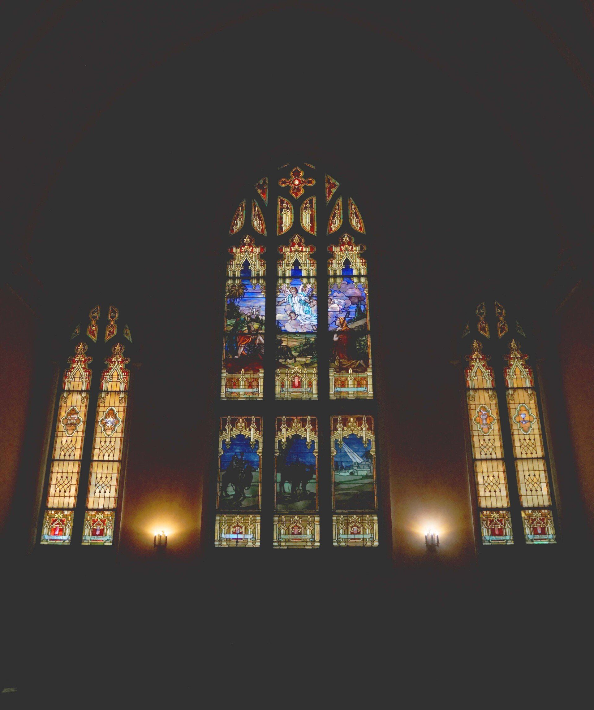 Three stained glass windows in a dark room. The central window is larger and features blue hues.