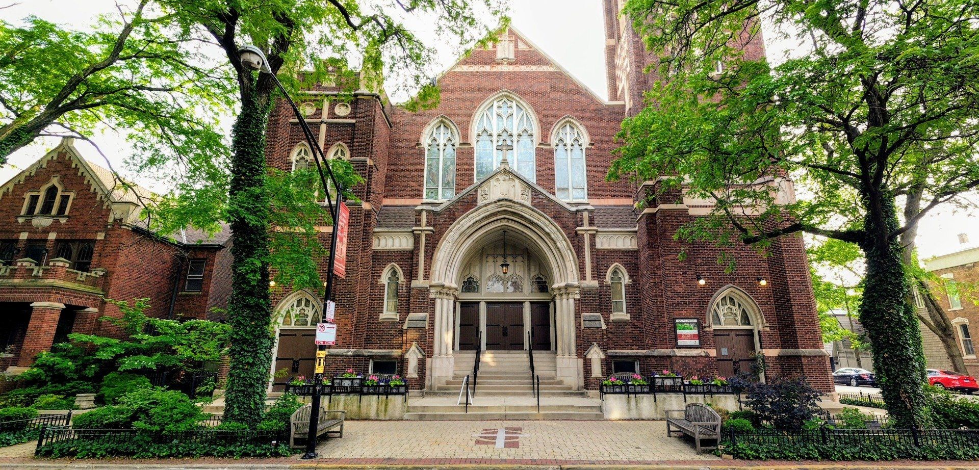 Red brick church with arched doorway and stained glass, surrounded by trees and greenery.