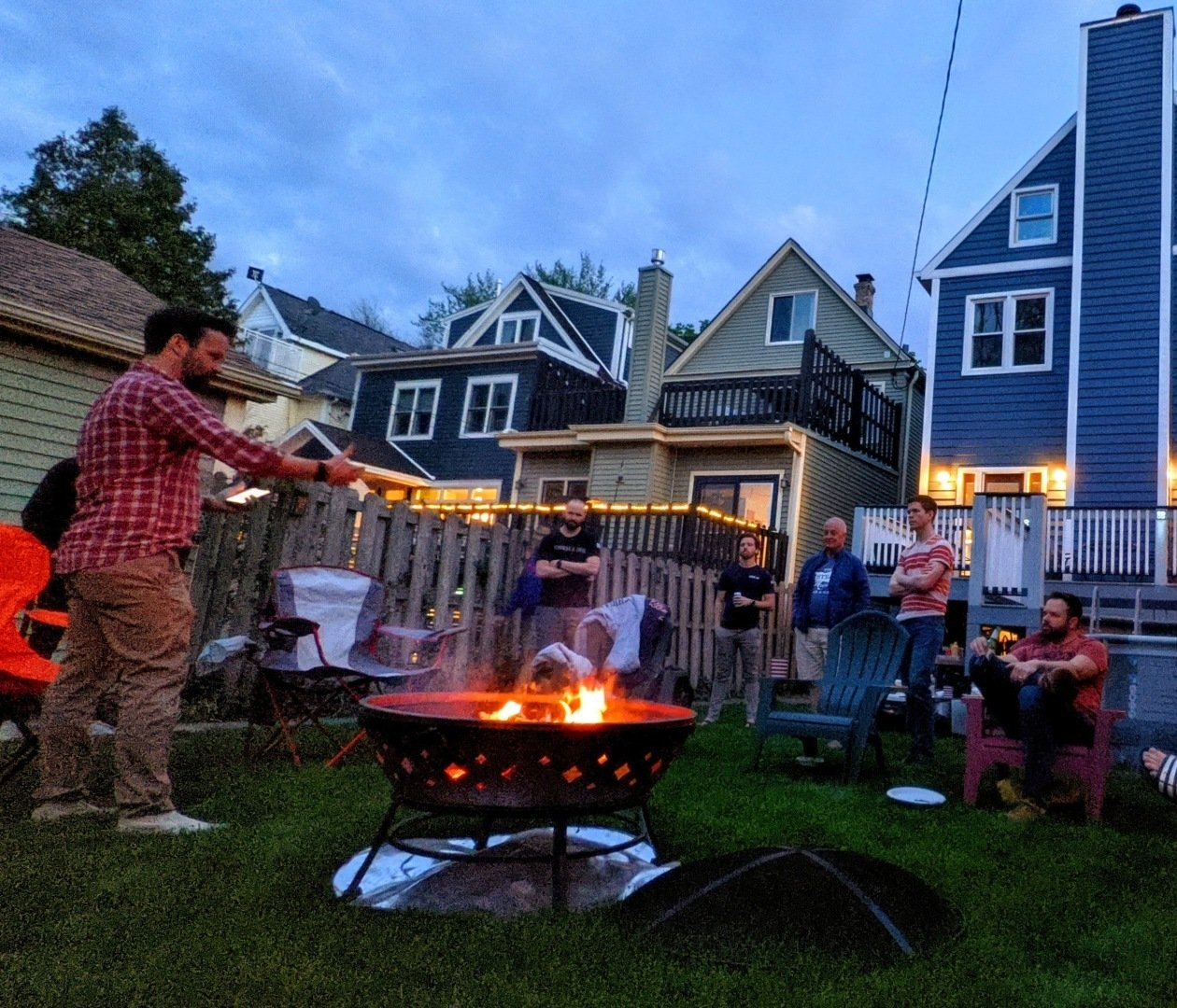 People gather around a fire pit in a backyard, roasting food at dusk.