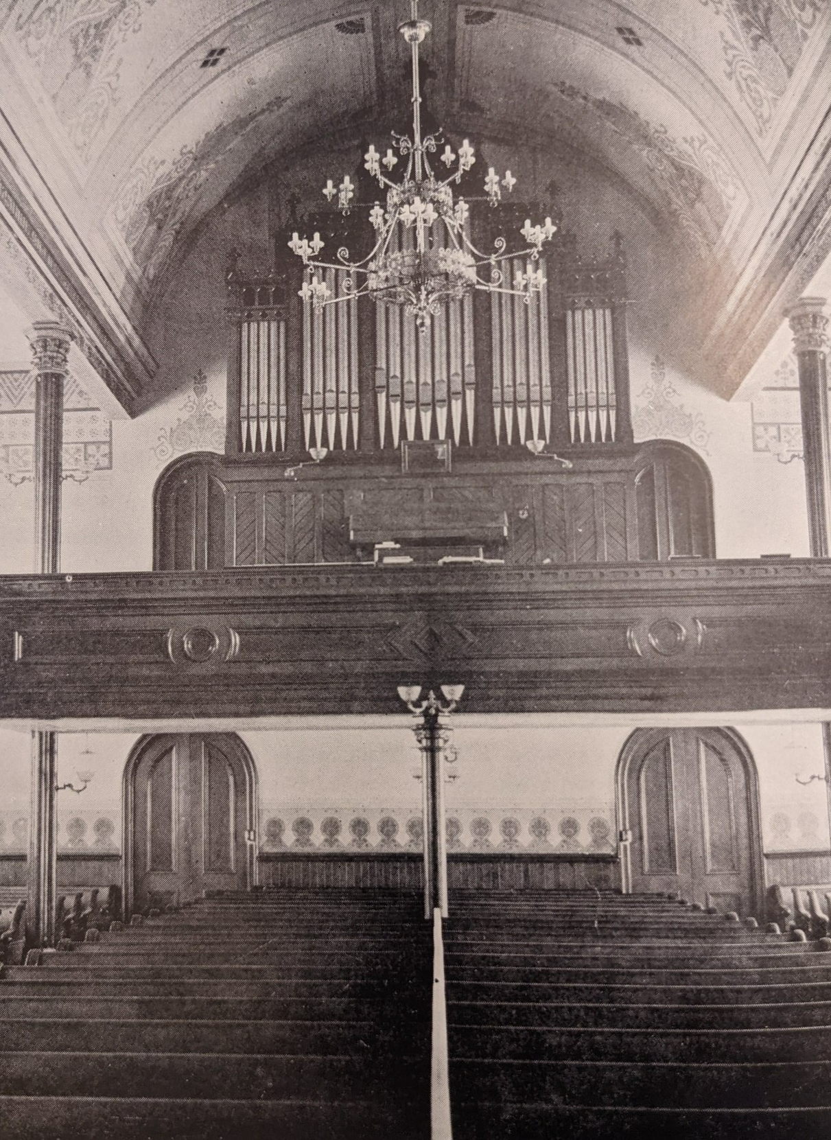 Interior view of a church. Organ pipes behind a wooden balcony. Rows of pews face the front. Arched ceiling with chandelier.