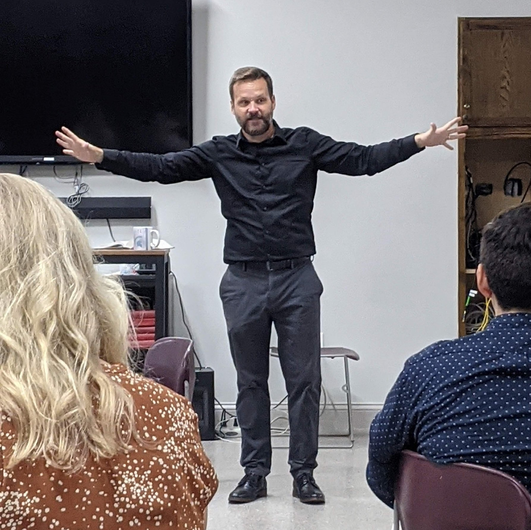 Man in black shirt with arms outstretched, giving a presentation to an audience in a room.