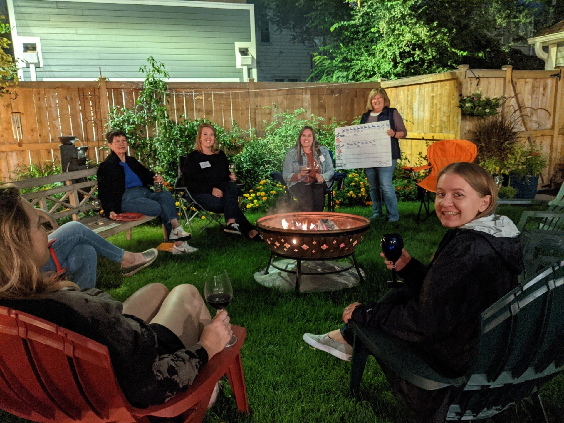 Group of people gathered around a fire pit in a backyard, some holding drinks, one holding a sign.